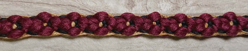 a braid with gold and black borders to either side of  five-petalled flowers that have black and gold centres. 