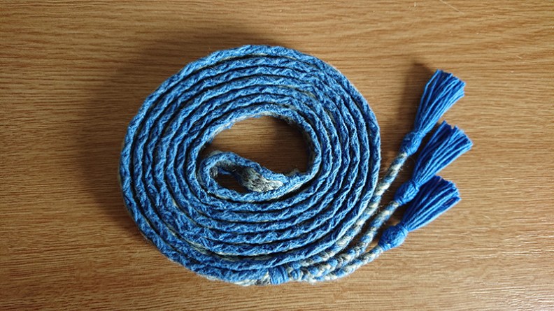 Tablet woven band with blue borders, coiled on itself, with three blue tassels to the right of the coiled band.